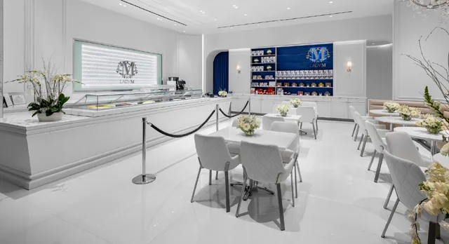 Interior view of Lady M at Tysons Galleria featuring a pristine white color scheme with elegant display cases showcasing cakes on the left and an illuminated Baccarat tile display with LADY M's logo. A distinctive blue accent wall with product displays adorns the back of the space, while white quilted chairs and small tables with fresh flower arrangements provide comfortable seating for guests.