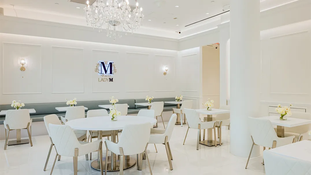 Elegant dining area of the Lady M Bellevue boutique featuring a luxurious crystal chandelier, pristine white walls with decorative paneling, and the Lady M logo displayed prominently. The space includes white tables with white quilted chairs, fresh flower arrangements, and a soft gray banquette seating along the wall, all creating a sophisticated atmosphere for guests.