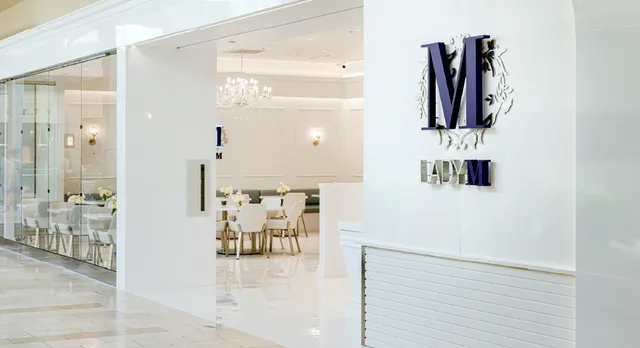 The entrance of Lady M's Bellevue boutique showing a sleek white exterior with glass windows and the elegant Lady M logo prominently displayed on the wall. Through the doorway, you can see the sophisticated interior with white tables, quilted chairs, a crystal chandelier, and the signature white and blue color scheme that creates an upscale atmosphere for guests.