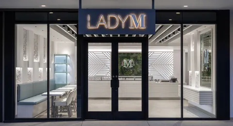 Lady M Sawtelle storefront with gold sign, glass doors, white interior, blue seats, and moss logo.