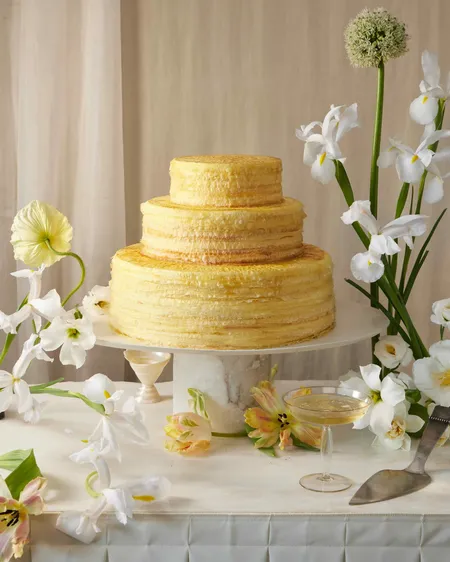 Three-tiered Lady M Signature Mille Crêpes cake with white and yellow flowers and champagne.