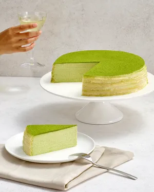 A hand holds a drink over a sliced Lady M Green Tea Mille Crêpes and a slice on a plate.
