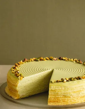 A Pistachio Mille Crêpes cake with a slice served on a plate, topped with chopped pistachios.