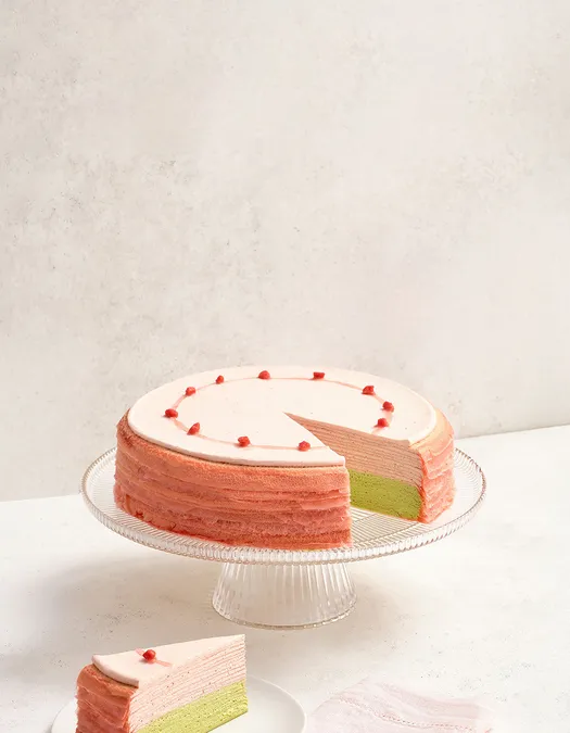 Lady M's Strawberry Matcha Mille Crêpes on a glass cake stand with a stone surface and background.