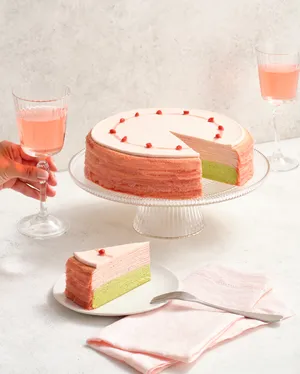 Lady M's Strawberry Matcha Mille Crêpes on a glass cake stand with a slice in front and a hand holding a rosé wine glass.