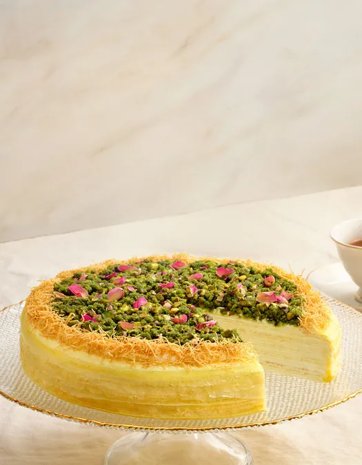 Lady M’s Baklava Mille Crêpes on glass stand, slice plated on beige tablecloth with tea.