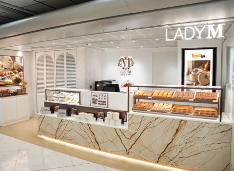 View of the sleek Lady M New York cake boutique storefront, featuring a marble counter, diverse rows of pastries, and a large neon logo.