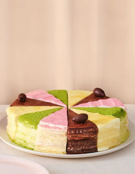 A round, multi-layered crepe cake sliced into even pieces, each topped with a different flavored powder in pastel colors—green, pink, yellow, and chocolate brown—and garnished with small chocolate curls, displayed on a white plate against a neutral background