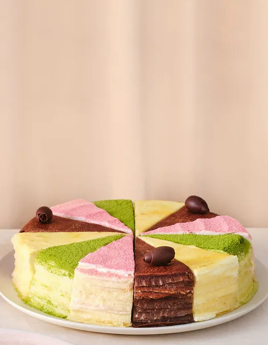 A round, multi-layered crepe cake sliced into even pieces, each topped with a different flavored powder in pastel colors—green, pink, yellow, and chocolate brown—and garnished with small chocolate curls, displayed on a white plate against a neutral background