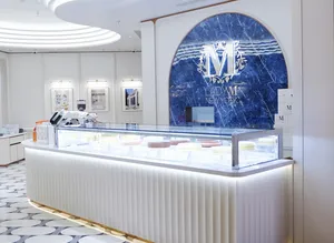 Bright, modern dessert boutique interior with a glowing curved ceiling design, patterned black-and-white tile floor, and a glass display counter showcasing cakes. A large blue marble wall behind the counter features the “Lady M New York” logo in silver lettering.