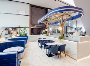 Elegant café interior with curved blue booth seating, marble floors and counters, and sculptural gold-and-blue canopy lighting above the service bar. Small dining tables and chairs are arranged throughout the bright, modern space, with “Lady M New York” signage displayed on the wall in the background.