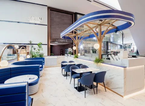 Elegant café interior with curved blue booth seating, marble floors and counters, and sculptural gold-and-blue canopy lighting above the service bar. Small dining tables and chairs are arranged throughout the bright, modern space, with “Lady M New York” signage displayed on the wall in the background.