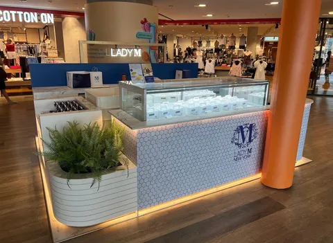 A high-angle, medium shot shows a Lady M New York pop-up boutique in a brightly lit shopping mall. The boutique is a rectangular kiosk with white and grey marble countertops and a white honeycomb-patterned base. A glass display case sits on top of the kiosk, filled with white Lady M gift boxes. A large, green fern is planted in a white, slatted planter at the front of the kiosk. The Lady M New York logo is prominently displayed on the side of the kiosk. In the background, other mall shops and shoppers can be seen. The lighting is bright and even, highlighting the clean and modern design of the boutique.