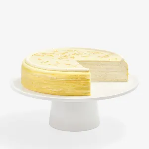 A multi-layered Osmanthus Mille Crêpes cake with one slice removed, displayed on a minimalist white ceramic cake stand against a solid white background. The cake features dozens of paper-thin crêpe layers alternated with light pastry cream. Its smooth, pale-cream glazed top is bordered by a neat ring of piped cream and delicately sprinkled with golden-yellow osmanthus flower petals.
