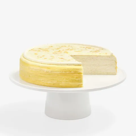A multi-layered Osmanthus Mille Crêpes cake with one slice removed, displayed on a minimalist white ceramic cake stand against a solid white background. The cake features dozens of paper-thin crêpe layers alternated with light pastry cream. Its smooth, pale-cream glazed top is bordered by a neat ring of piped cream and delicately sprinkled with golden-yellow osmanthus flower petals.