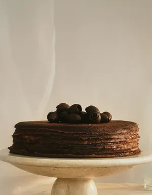 A wide banner featuring a chocolate mille crêpes cake on a marble stand, positioned on the right side with ample negative space on the left. The cake is topped with chocolate shavings against a soft cream backdrop.