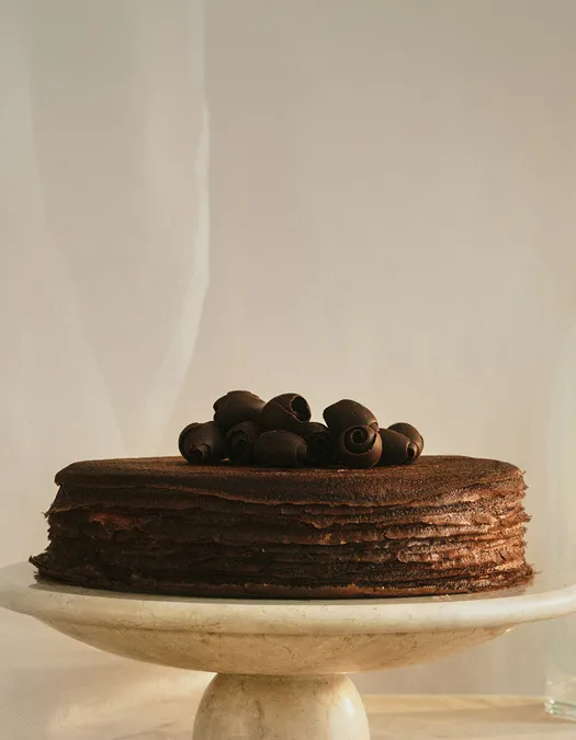 A wide banner featuring a chocolate mille crêpes cake on a marble stand, positioned on the right side with ample negative space on the left. The cake is topped with chocolate shavings against a soft cream backdrop.