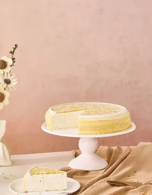 A wide desktop banner featuring an osmanthus mille crêpes cake with a slice missing on a white stand, positioned on the left side. The cake features a gold-flecked flower glaze, set against a textured dusty pink backdrop with empty negative space on the right.