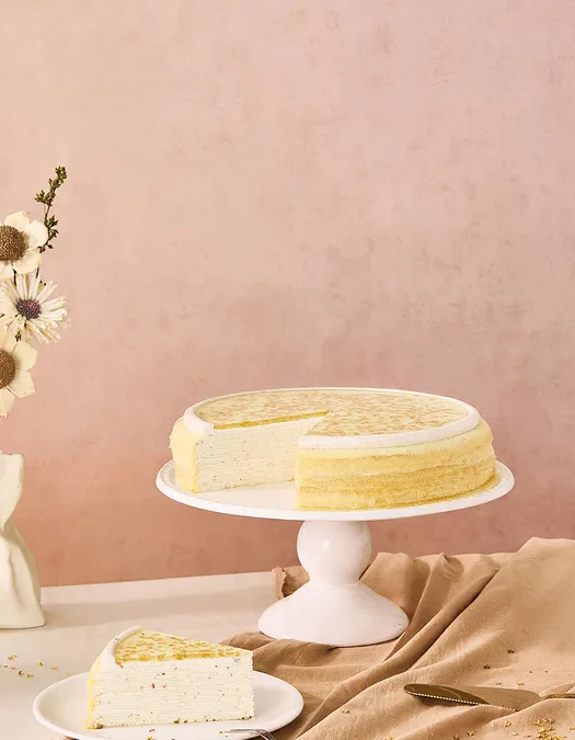 A wide desktop banner featuring an osmanthus mille crêpes cake with a slice missing on a white stand, positioned on the left side. The cake features a gold-flecked flower glaze, set against a textured dusty pink backdrop with empty negative space on the right.