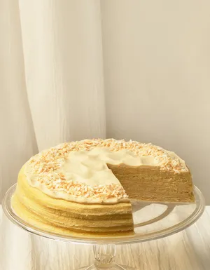A wide desktop banner showing a tres leches mille crêpes cake with a slice missing on a clear glass stand. The cake is positioned on the left, showcasing its layered cream and toasted coconut topping, with ample empty space on the right against a white linen background.