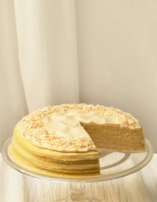 A wide desktop banner showing a tres leches mille crêpes cake with a slice missing on a clear glass stand. The cake is positioned on the left, showcasing its layered cream and toasted coconut topping, with ample empty space on the right against a white linen background.
