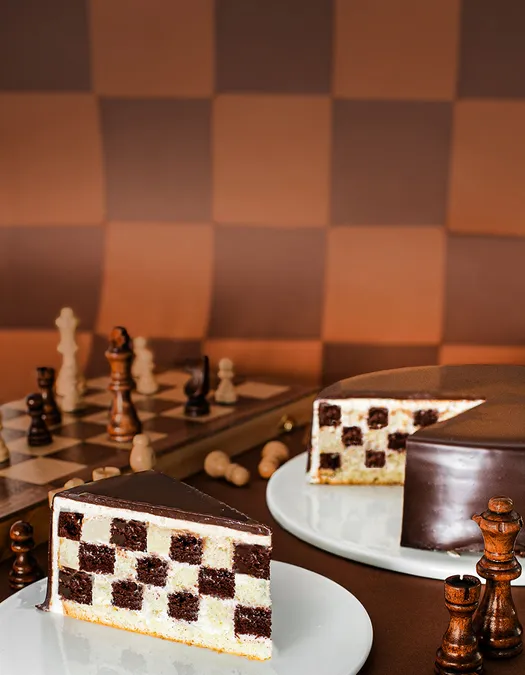 A wide desktop banner with a large solid dark brown block of negative space on the left. The right side features a chocolate-glazed checkerboard cake with a cut slice, set alongside a wooden chessboard and chess pieces.