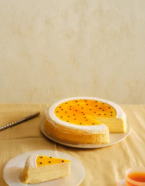 A wide desktop banner featuring a passion fruit mille crêpes cake with a bright yellow seed-flecked glaze, positioned on the right side with a slice missing. The left side provides open negative space against a warm, light beige background.