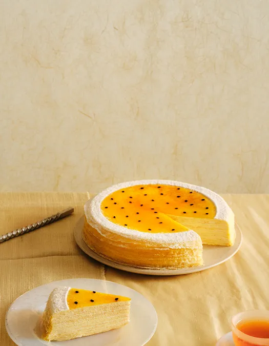 A wide desktop banner featuring a passion fruit mille crêpes cake with a bright yellow seed-flecked glaze, positioned on the right side with a slice missing. The left side provides open negative space against a warm, light beige background.