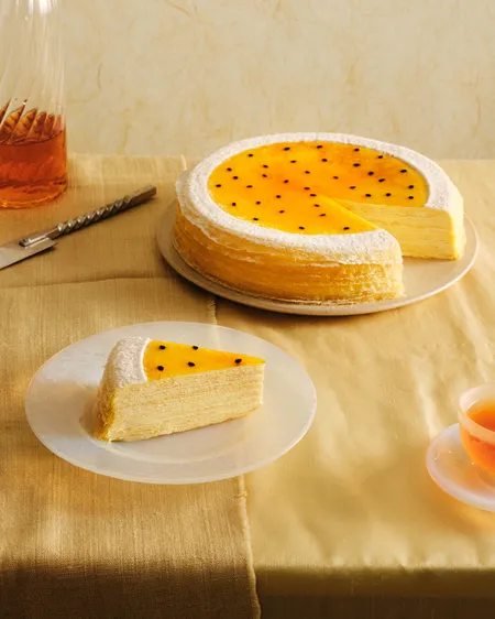 A passion fruit mille crêpes cake on a beige linen tablecloth, with one slice served on a clear plate in the foreground. The cake is topped with a vibrant yellow passion fruit glaze, black seeds, and a powdered sugar border.