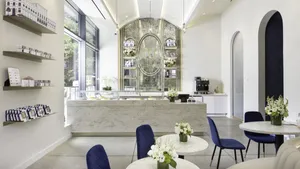 The bright, elegant interior of the Lady M Columbus Circle boutique, featuring a marble counter, blue velvet chairs, and a grand arched mirror behind the bar.