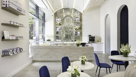 The bright, elegant interior of the Lady M Columbus Circle boutique, featuring a marble counter, blue velvet chairs, and a grand arched mirror behind the bar.
