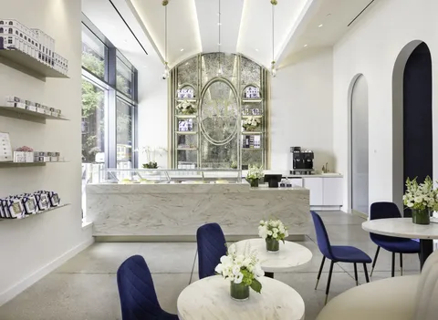 The bright, elegant interior of the Lady M Columbus Circle boutique, featuring a marble counter, blue velvet chairs, and a grand arched mirror behind the bar.