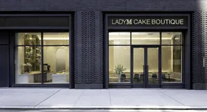 Exterior view of the modern Lady M Cake Boutique storefront with an illuminated gold sign reading 'LADY M CAKE BOUTIQUE' and large glass windows showing a brightly lit interior with marble counters and seating.