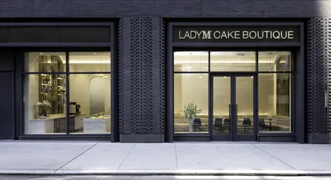 Exterior view of the modern Lady M Cake Boutique storefront with an illuminated gold sign reading 'LADY M CAKE BOUTIQUE' and large glass windows showing a brightly lit interior with marble counters and seating.
