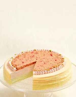 A layered Mille Crêpes cake on a clear glass stand with a slice removed, showing light green and cream-colored layers. The top features a textured pink crumb topping, a piped frosting border, and small green garnishes.