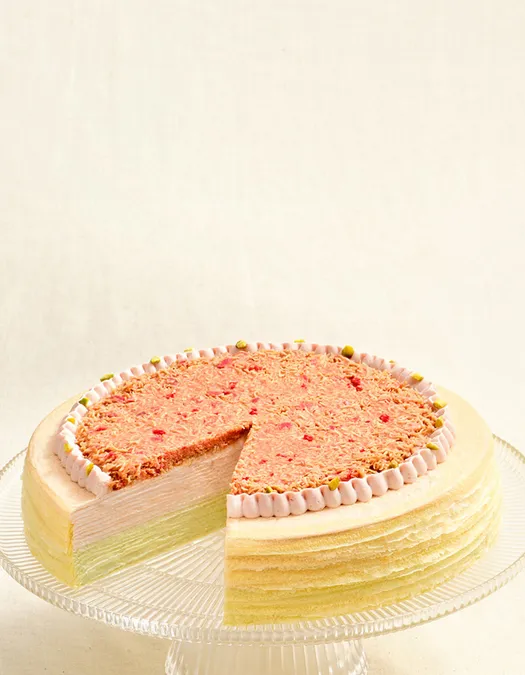 A layered Mille Crêpes cake on a clear glass stand with a slice removed, showing light green and cream-colored layers. The top features a textured pink crumb topping, a piped frosting border, and small green garnishes.