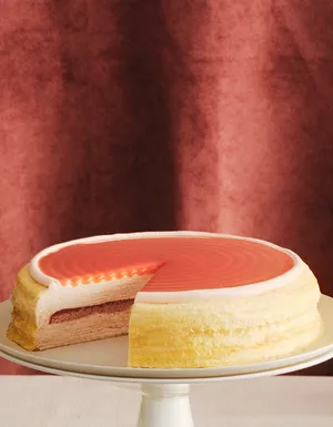 A layered Mille Crêpes cake on a stand with a slice removed, revealing cream-colored layers and a darker pink center layer. The cake is topped with a smooth, vibrant reddish glaze against a dark textured burgundy background.