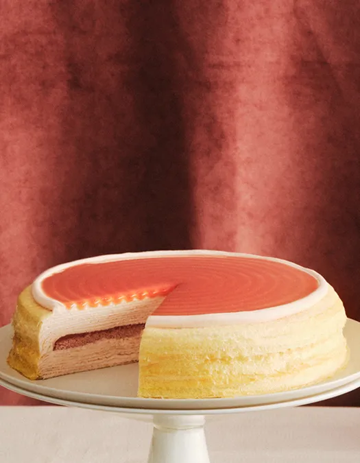 A layered Mille Crêpes cake on a stand with a slice removed, revealing cream-colored layers and a darker pink center layer. The cake is topped with a smooth, vibrant reddish glaze against a dark textured burgundy background.