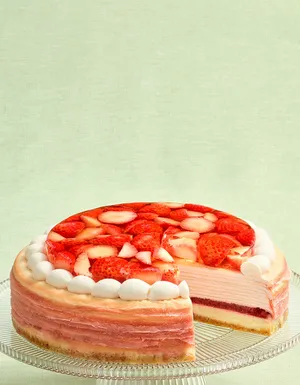 A pink layered Mille Crêpes cake on a clear glass stand with a slice removed, revealing a dark red fruit layer and cream layers inside. The top is covered with sliced red strawberries in a clear glaze and bordered by white frosting dollops against a light green background.