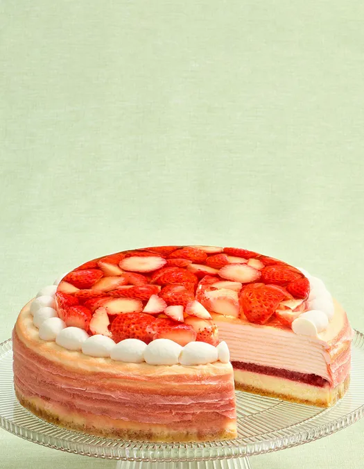 A pink layered Mille Crêpes cake on a clear glass stand with a slice removed, revealing a dark red fruit layer and cream layers inside. The top is covered with sliced red strawberries in a clear glaze and bordered by white frosting dollops against a light green background.
