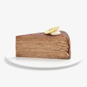 A slice of layered chocolate Mille Crêpes cake served on a plain white plate against a white background. The side shows dozens of thin brown crepe and light cream layers, topped with a dollop of whipped cream and a banana slice garnish.