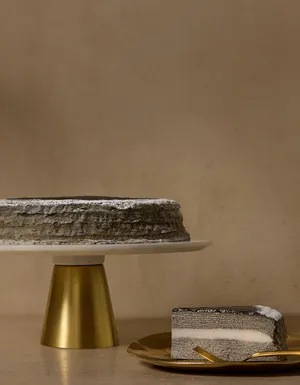 A grey layered Mille Crêpes cake on a gold-and-white pedestal stand alongside a slice served on a textured gold plate with a gold fork. The cut slice reveals thin charcoal-grey crepe layers with a thick, smooth white center layer, topped with a dark glaze and a light dusting of white powder against a warm beige backdrop.