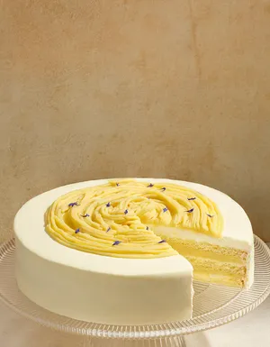 A white frosted layer cake on a clear ribbed glass stand with a slice removed, revealing light yellow sponge cake and cream layers inside. The top is decorated with swirled yellow cream piping accented with tiny purple flower petals, set against a soft cream background.