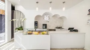 Interior view of a bright, modern Lady M boutique featuring a white fluted counter with a glass display case filled with whole cakes. A potted white orchid rests on the left, three pendant lights hang above, and a silver Lady M logo is mounted inside an arched stone alcove behind the counter, all set over a speckled terrazzo floor.