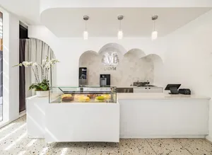 Interior view of a bright, modern Lady M boutique featuring a white fluted counter with a glass display case filled with whole cakes. A potted white orchid rests on the left, three pendant lights hang above, and a silver Lady M logo is mounted inside an arched stone alcove behind the counter, all set over a speckled terrazzo floor.