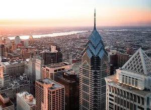 Aerial view of the Philadelphia skyline at sunset, featuring One Liberty Place and City Hall glowing in warm sunlight.