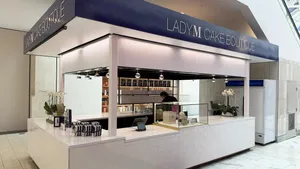 An indoor Lady M Cake Boutique kiosk featuring white tiled counters and a dark blue top banner reading 'LADY M CAKE BOUTIQUE'. The white counter displays a glass case, POS terminal, potted white orchids, and gift boxes, with organized shelving visible inside.
