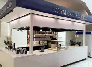 An indoor Lady M Cake Boutique kiosk featuring white tiled counters and a dark blue top banner reading 'LADY M CAKE BOUTIQUE'. The white counter displays a glass case, POS terminal, potted white orchids, and gift boxes, with organized shelving visible inside.