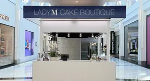 A front view of the Lady M Cake Boutique kiosk inside a bright shopping mall corridor under a dark blue sign reading 'LADY M CAKE BOUTIQUE'. The white tiled counter holds potted white orchids and a checkout terminal, situated in the concourse between Nespresso and Under Armour