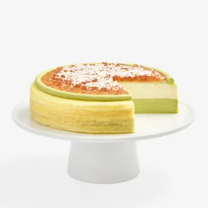 A whole Mille Crêpes cake on a white pedestal stand with a slice cut out, showcasing light green bottom layers and cream top layers. The surface features an amber-colored jelly topping dusted with fine white flakes and encircled by a green frosting ring.